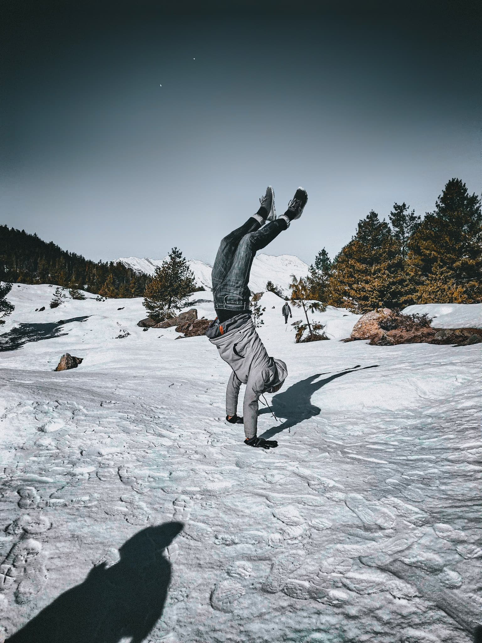 A trekker performs a handstand on a vast snow-covered glacier field with pine trees behind