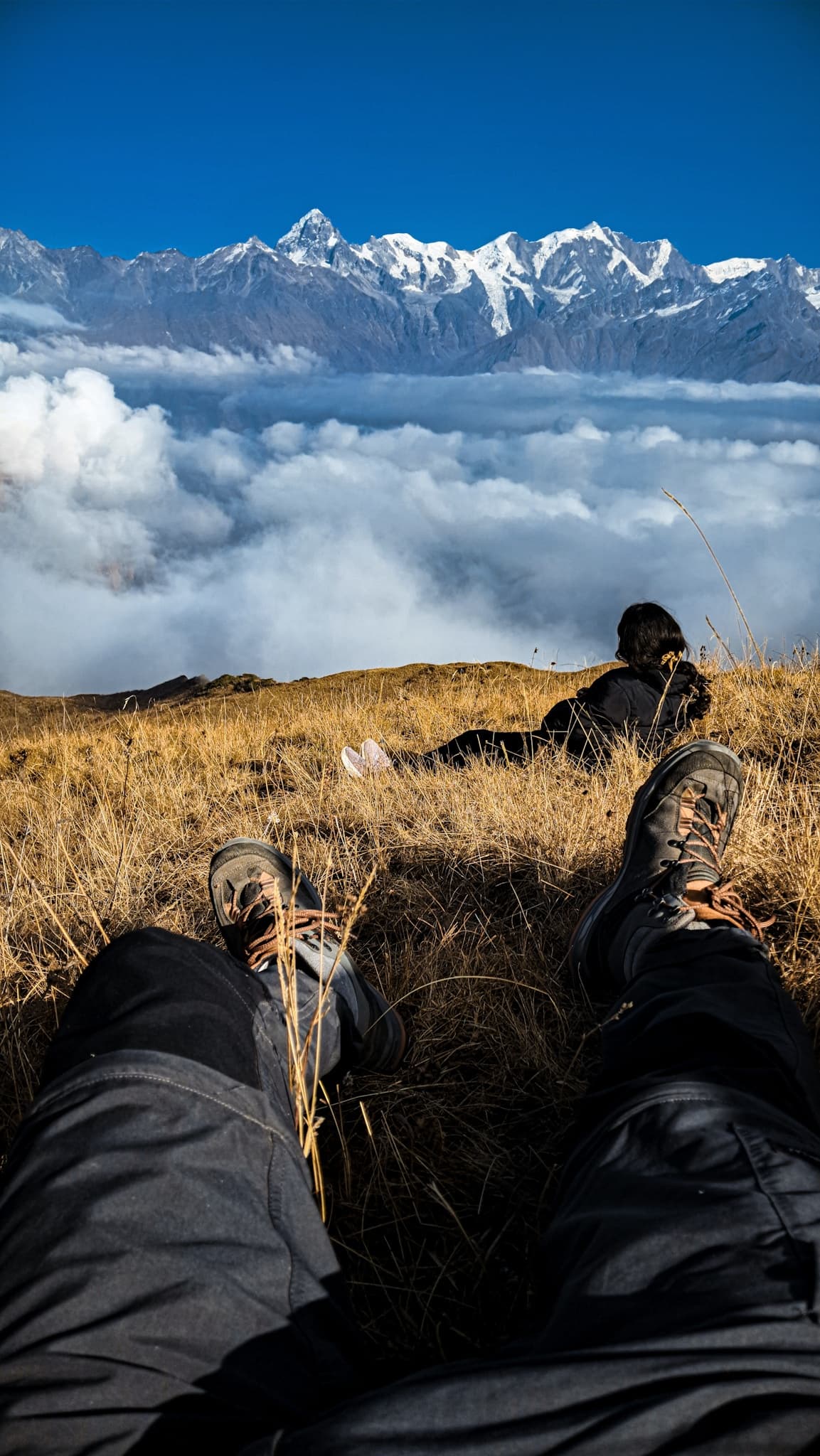 Two trekkers lying in alpine grass facing a panorama of snow-capped Himalayan peaks