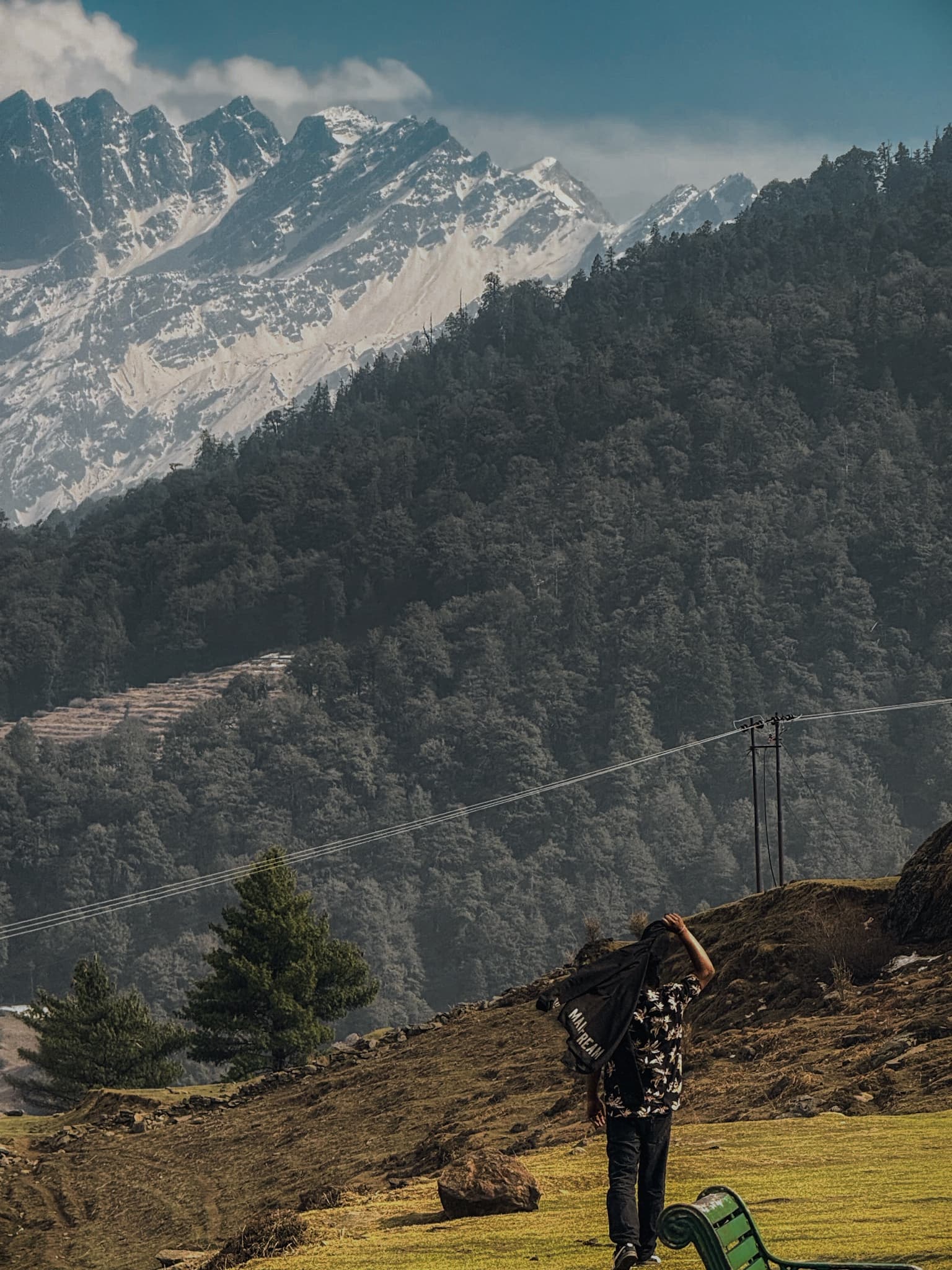 A solo traveller with a backpack stands before lush green meadows and massive snow-streaked peaks
